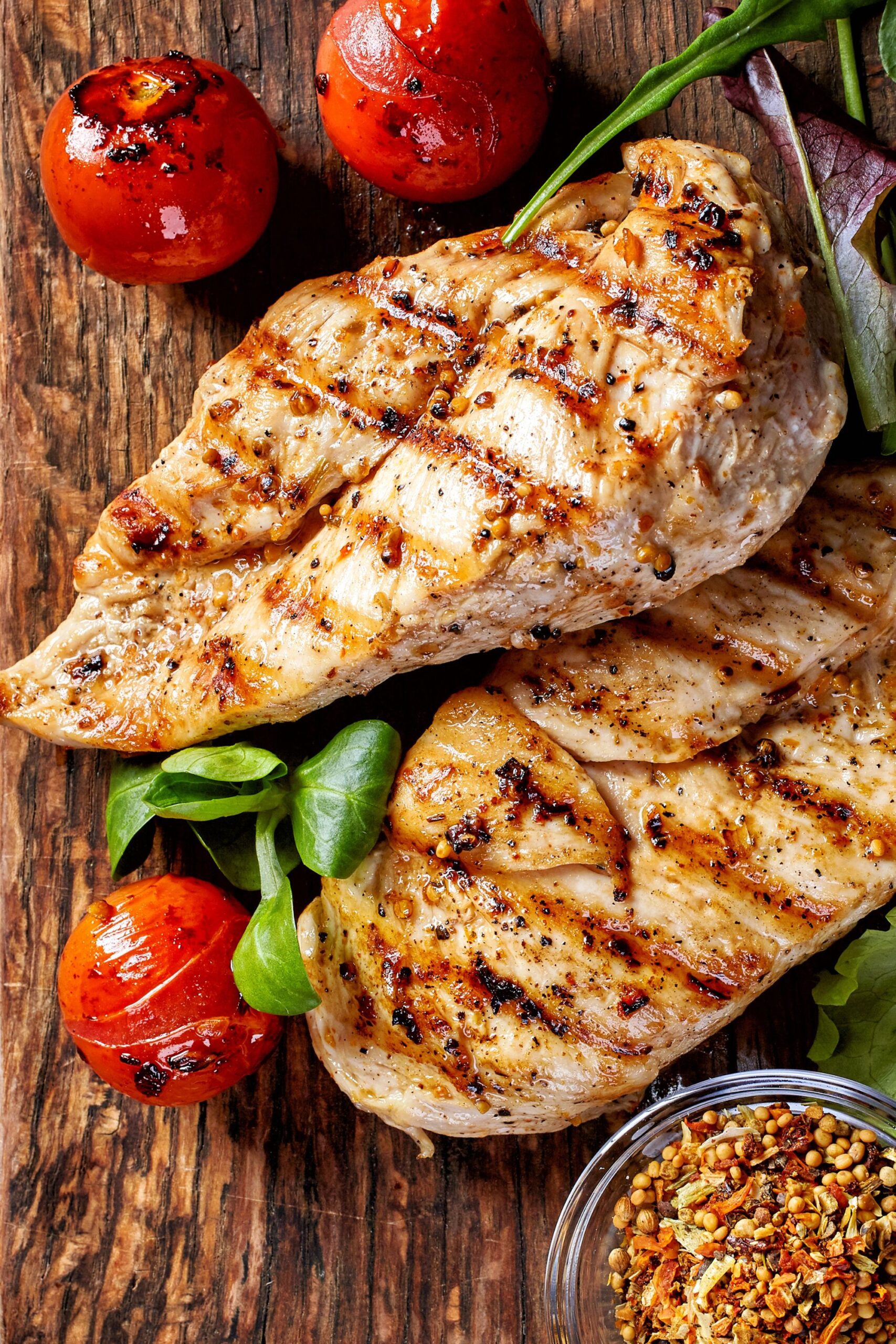 two pieces of grilled chicken breasts next to grilled tomatoes and a bowl of spices