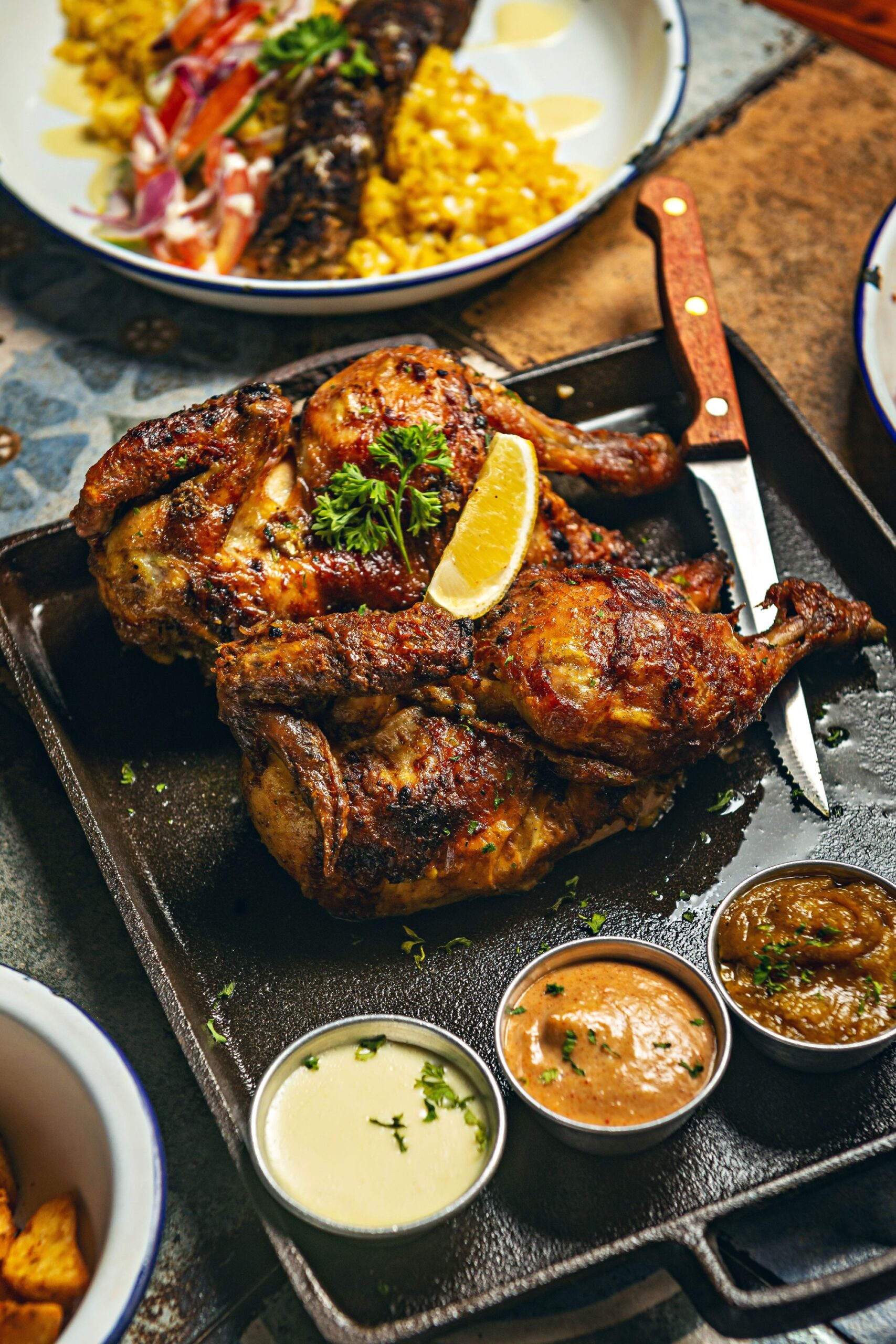 Whole grilled chicken on a black plate with a lemon on top and little sauces on the side.